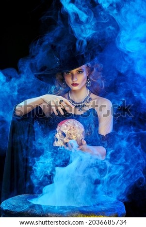 Halloween magic. A beautiful young witch in a hat and elegant dress conjures with a skull over a cauldron surrounded by magical smoke.