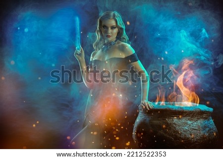 Halloween magic. A beautiful young witch in an elegant black off-the-shoulder dress conjures at night over a cauldron with a magic wand. Copy space.