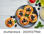 Halloween mini spider pizzas made from.pizza crust,pizza sauce,olive oil,mozzarella cheeses and black olives on plate with white wood table background.Art food idea per kids halloween party.Copy space