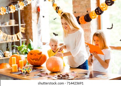 Halloween mood. Blonde family of mother and two siblings are preparing for party, cutting decorations, making jackolantern, desktop with candles, candies, fall leaves, little black bats on windows