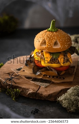 Halloween party burger in shape of scary pumpkin on natural wooden board on dark background. Scary Halloween food concept. Creative Halloween cheeseburger with cheese, cutlet, tomato, pepper, salad.