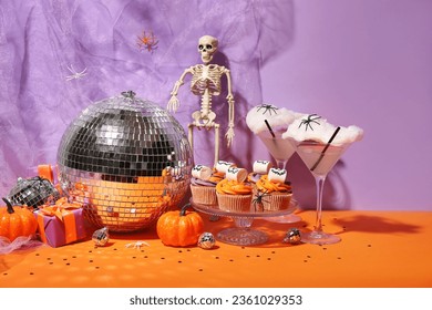 Halloween party composition with cupcakes, disco ball, drinks and decorations on orange table against purple background - Shutterstock ID 2361029353
