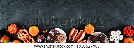 Halloween party food bottom border over a black stone banner background with copy space. Top view. Spooky mummy pizzas, finger hot dogs, caramel apples, cupcakes, donuts, cookies and candy. 