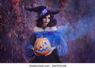 Halloween portrait fantasy woman witch. Girl sorceress in vintage purple costume medieval dress, tall cone hat. Magician is holding orange Jack-o'-lantern pumpkin with blue smoke. autumn background.