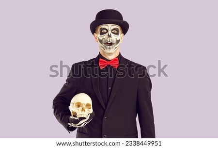 Halloween. Portrait of man with skeleton makeup who is looking at you with surprised and shocked expression. Man in black suit and hat holds decorative skull, isolated on light lilac background.