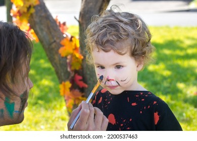 Halloween Preparation To Party. Finishing Costume With Makeup, Dad Painting On Face Of Son With Aqua Paint, Face Painting For Blond Curly Boy. Trick Or Treat. Family Celebration In Carnival Costumes