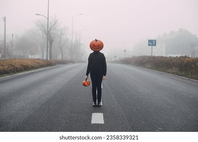 Halloween. Pumpkinhead. A man with a pumpkin on his head stands in the middle of a foggy street. Holds a jack-o'-lantern in his hand - Powered by Shutterstock