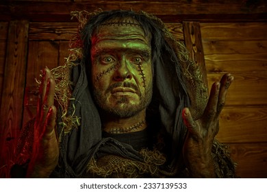 Halloween. A scary bearded man with scars on his face, dressed in burlap clothes, looking at the camera with scary eyes, against a wooden house. Horror fantasy novel. Frankenstein monster.