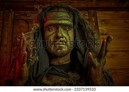 Halloween. A scary bearded man with scars on his face, dressed in burlap clothes, looking at the camera with scary eyes, against a wooden house. Horror fantasy novel. Frankenstein monster.