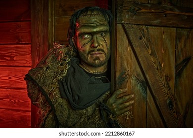 Halloween. A scary man with scars on his face and neck, dressed in burlap clothes, hides behind the door of a wooden hut. Frankenstein monster. Gothic novel, horror. 