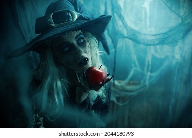 Halloween. A terrible wicked witch holds a poisoned apple in her hands, standing in her lair among the cobwebs. Evil black magic.