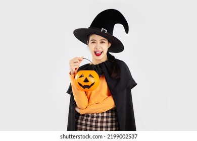 Halloween theme, young pretty asian woman in black halloween costume wearing witch hat holding orange pumpkin bucket posing on white background.