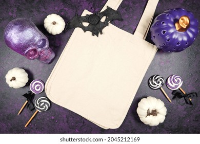 Halloween Trick Or Treat Canvas Tote Bag Mockup With Purple Skull And White Pumpkins, Lollipops, Candy, And Bat, On A Textured Purple Background. Product Mock Up With Negative Copy Space.