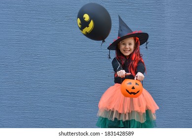 Halloween Trick Or Treat. Portrait Smiling Girl In Witch Hat With Balloon And Pumpkin Bucket For Sweets Against A Blue Wall . Copy Space For Text