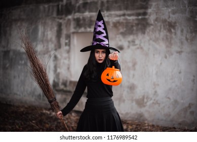 Halloween witch with a broomstick. Beautiful young woman in witch costume outdoors. Beautiful young woman in witches hat and holding pumpkin.