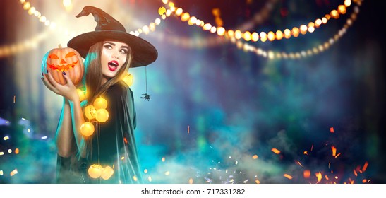 Halloween Witch with a carved Pumpkin and magic lights in a dark forest. Beautiful young surprised woman in witches hat and costume holding pumpkin. Wide Halloween party art design