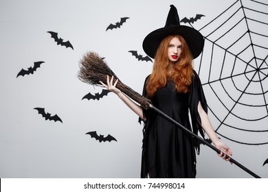 Halloween Witch Concept - Portrait of beautiful young witch with broomstick over grey wall with bat and spider web background.