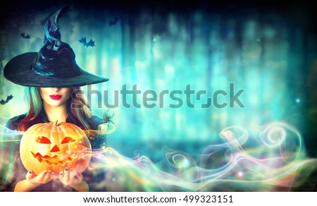 Halloween Witch with a magic Pumpkin in a dark forest. Beautiful young woman in witches hat and costume holding carved pumpkin. Wide Halloween art design