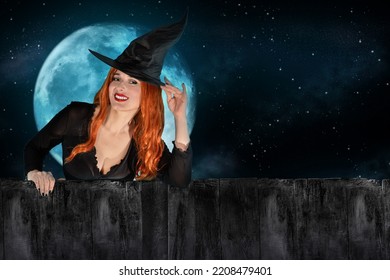 Halloween Witch peeking out behind a fence or wooden wall. Female wizard fairy character. All Saints' Day. Fantasy gothic red-haired sorceress girl dressed in black carnival costume. Enchantress woman