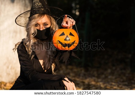 Halloween Witch with pumpkin. Beautiful young woman in a witch costume and black mask on the street near the house. To stop the pandemic COVID-19. Halloween decor.