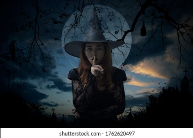 Halloween witch showing silence sign with finger standing over cross, church, crow, bat, birds, dead tree, full moon and sunset sky, Halloween mystery concept