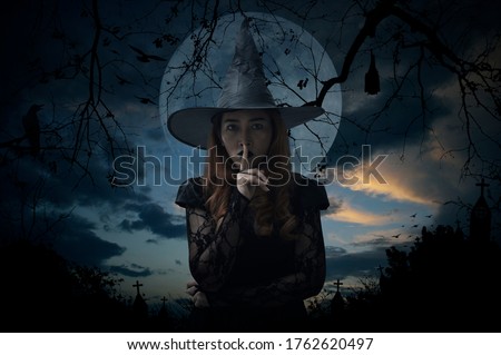 Halloween witch showing silence sign with finger standing over cross, church, crow, bat, birds, dead tree, full moon and sunset sky, Halloween mystery concept