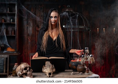 Halloween, Witch Use Magic Book And Cauldron Prepare Poison Or Love Potion. Black Magic Occult Female Wizard In Dark Room Cage Spider Web Human Skull. Necromancer Looking Camera