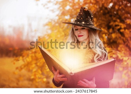 Halloween a woman with a magic spell book in a witch hat is conjuring, performing witchcraft over the autumn background of the forest.