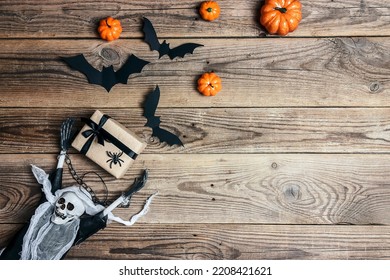 Halloween Wooden Background With Creepy Skeleton Holding A Gift, Pumpkins And Bats. Top View With Copy Space For Text.