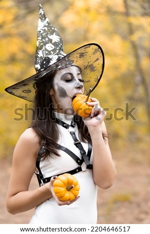 Halloween Young beautiful girl in a white dress with make-up on her face, in a witch's hat posing in the autumn forest. Skeleton face makeup. Bites a small pumpkin. The day of the Dead
