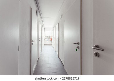 Hallway In A House With Many Solid White Doors That Have Round Handles Of Brushed Metal. One Door Has A Key In The Lock. At The End Of The Corridor There Are Pipes And A Window.