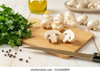 Halved raw champignon and kitchen knife on a cutting board, bunch of parsley and whole button mushrooms on a white wooden table. Cooking fresh white mushrooms. Vegetable protein. Front view. - Powered by Shutterstock