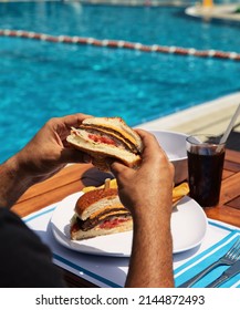 hamburger by the pool. The man by the pool is eating a hamburger. Burger with cola. burger on wooden plate. hamburger and swimming pool in summer. Poolside hamburger with ingredients showing.