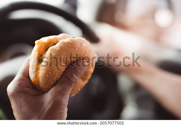 A\
hamburger in the hand of a chauffeur who is driving, Concept of\
travel activities eating fast food in the\
car.