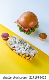 A Hamburger That Contains Lettuce, Tomato And Bologna, A Paper Dish Of French Fries With Alfredo Sauce And Two Tiny Bowl Of Sauces On A White And Yellow Background.