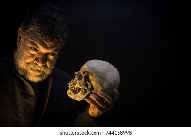 In A Hamlet Pose, A Bearded Crazy Old Man Looks Into A Skull's Eyes In Halloween.