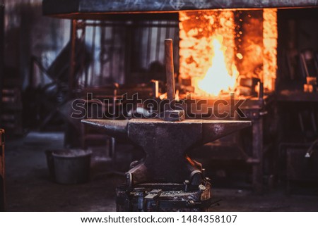 Hammer on anvil at dark blacksmith workshop with fire in stove at background.