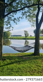 Hammock hanging between trees on pond shore with reflections in calm water and rural landscape. Relaxation and leisure concept