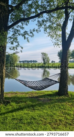 Hammock hanging between trees on pond shore with reflections in calm water and rural landscape. Relaxation and leisure concept