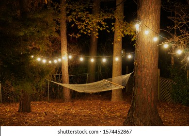 A Hammock Is Suspended Between Two Trees In A Backyard With Cafe Lights Strung Up; Romantic Nighttime Setting; Landscape View