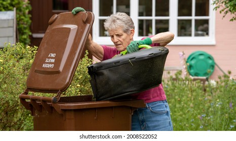 Hampshire, England, UK. 2021. Woman Emptying Grass Cuttings Into A Garden Waste Brown Wheelie Bin.