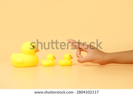 Hand about to flick two small rubber ducks beside a larger duck on a pastel background. Concept of leadership, influence, mentorship, growth and guiding the next generation.