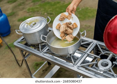Hand adding cooked pork pieces from a plate to a simmering pot on an outdoor gas stove, preparing food in a rustic setting