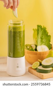 Hand adding a green apple slice into a portable blender filled with green detox smoothie, surrounded by fresh ingredients like cucumber, lime, and lettuce.