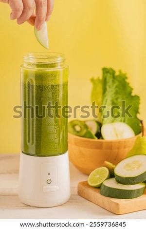 Hand adding a green apple slice into a portable blender filled with green detox smoothie, surrounded by fresh ingredients like cucumber, lime, and lettuce.