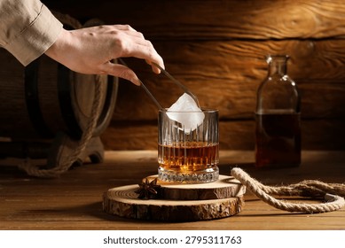 Hand adding ice cube to whiskey glass on wooden table with bottle and barrel in rustic setting, a hand uses tongs to place a large ice cube into a glass of whiskey, creating a refreshing beverage
