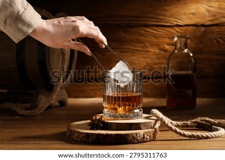 Hand adding ice cube to whiskey glass on wooden table with bottle and barrel in rustic setting, a hand uses tongs to place a large ice cube into a glass of whiskey, creating a refreshing beverage