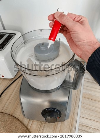Hand adding red food coloring into a food processor with cream mixture, preparation for cooking or baking in a kitchen, stainless steel appliance on a wooden surface