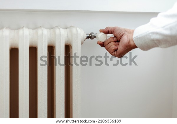 Hand adjusting the knob of heating radiator. Men's hand
adjusting thermostat valve of heating radiator in the bedroom.
Person's hand adjusts the temperature of the heater at home in
winter time. 