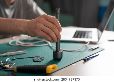 A hand adjusts a tool while standing at a worktable surrounded by electronic parts and a laptop.